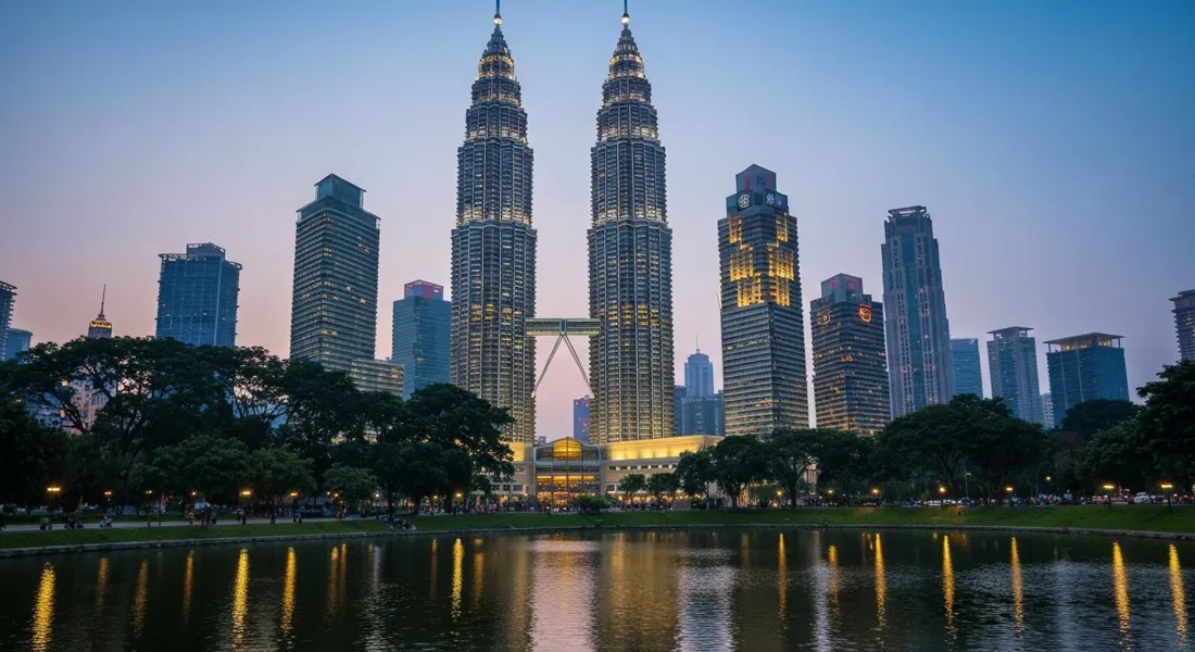 las Torres Gemelas Petronas elevándose sobre el Parque KLCC al atardecer, acero y vidrio atrapando la última luz dorada, con la línea de árboles y el lago en primer plano